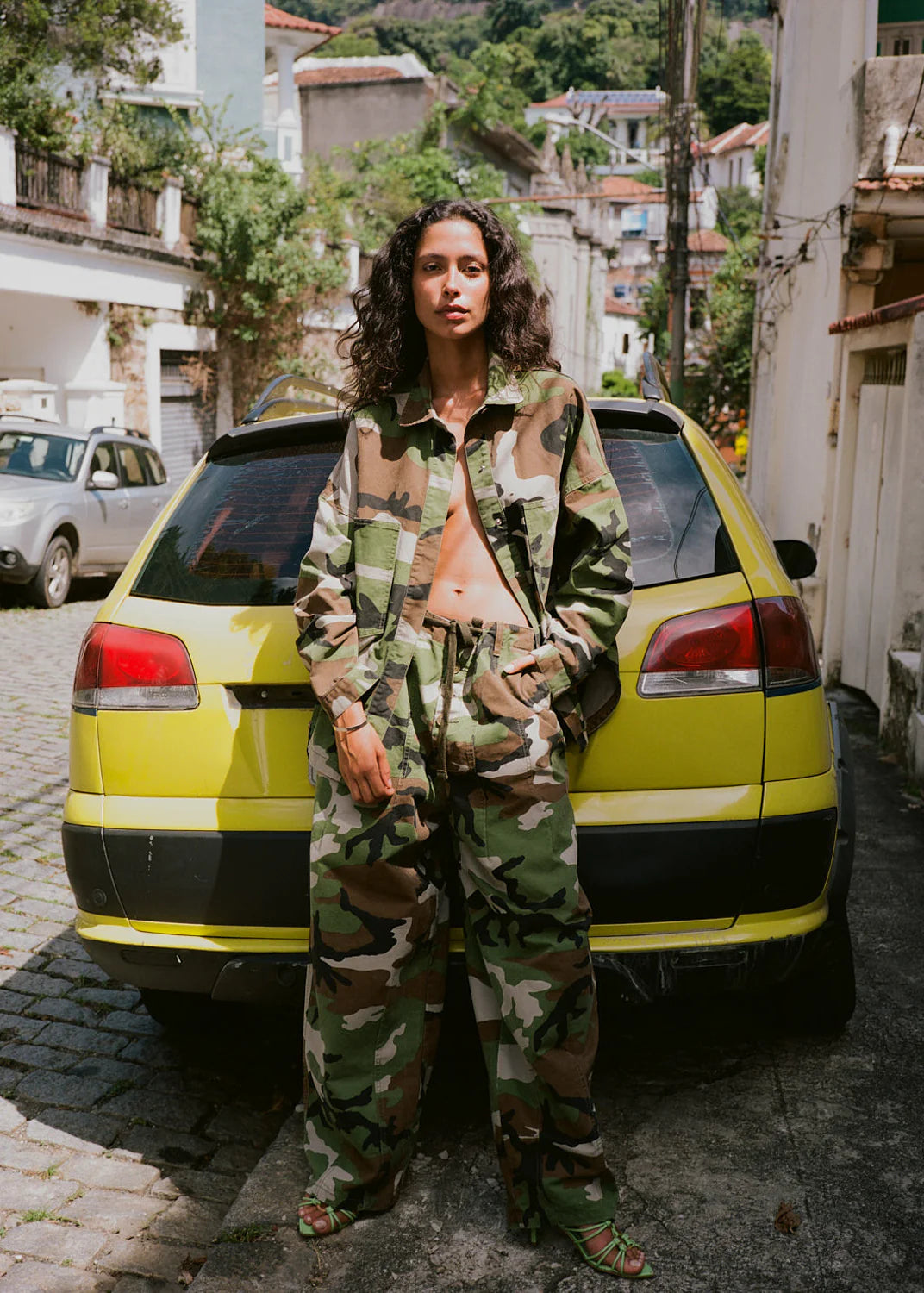 A woman stands outdoors in front of a bright yellow car on a cobblestone street, wearing a matching cammufare kinga csilla set. Military-style shirt and pants with draw cord and pockets