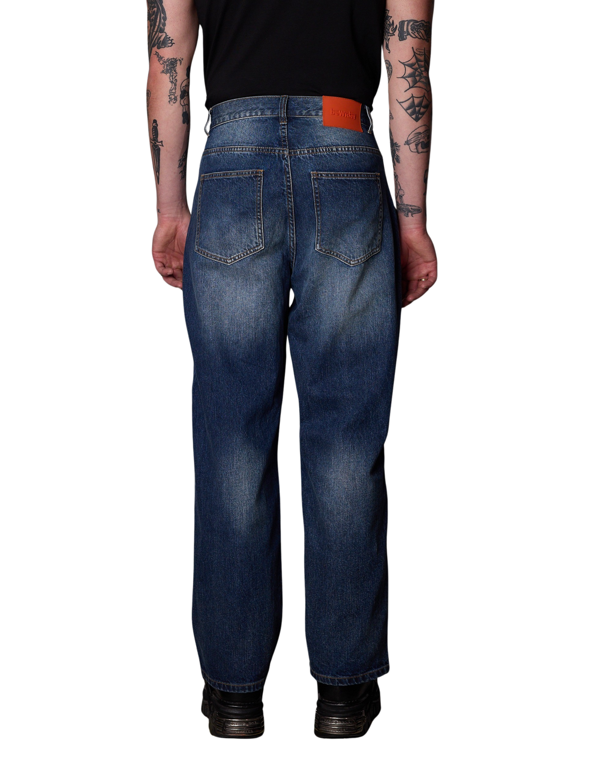 Rear view of a person wearing BeWider Super Loose Blue Dream jeans. Featuring the signature orange brand patch worn with a black shirt and black shoes, shown against a plain white background.