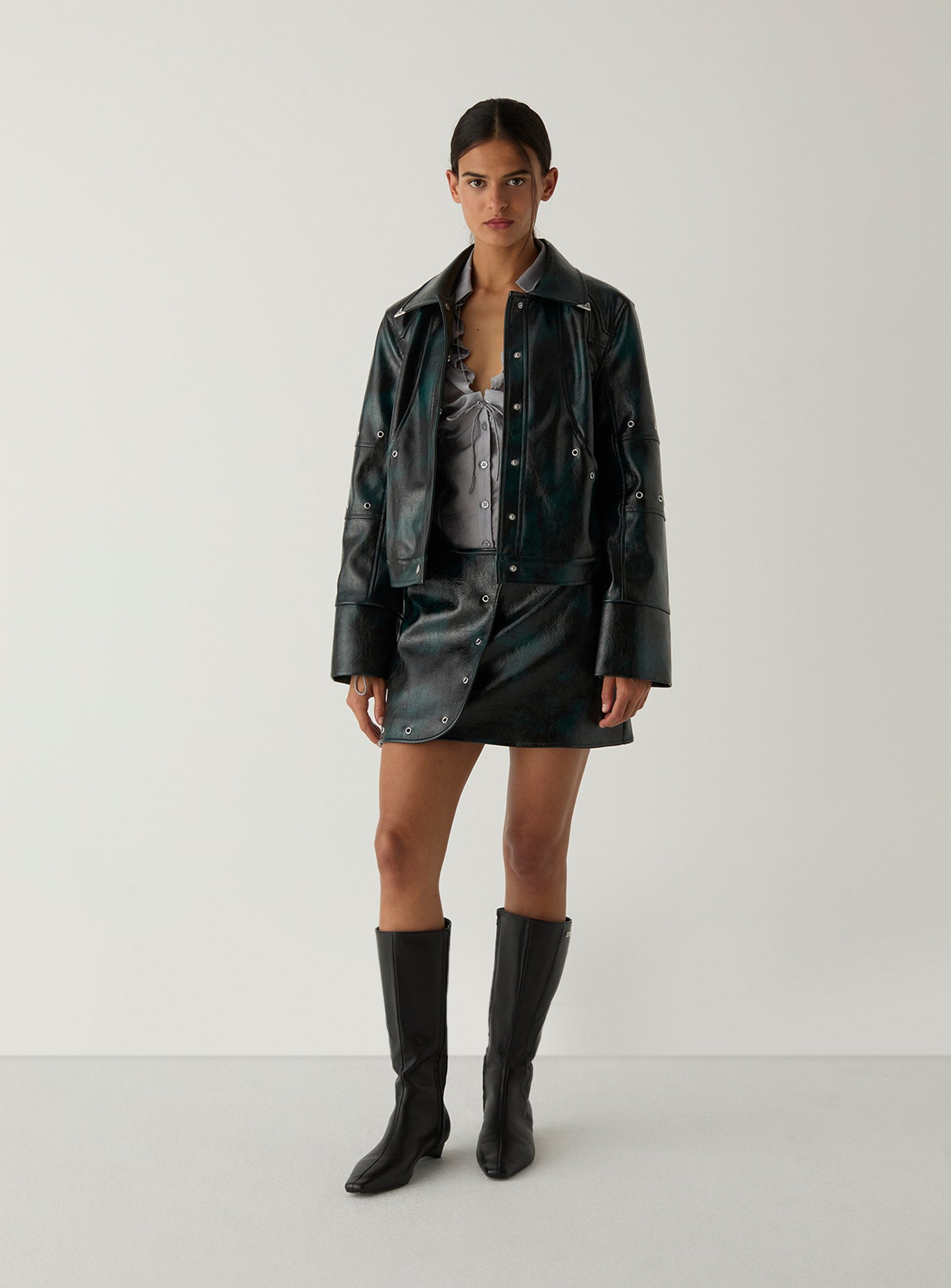 A model stands against a light gray studio background wearing a black faux-leather jacket, matching mini skirt, patterned top, and black knee-high boots.