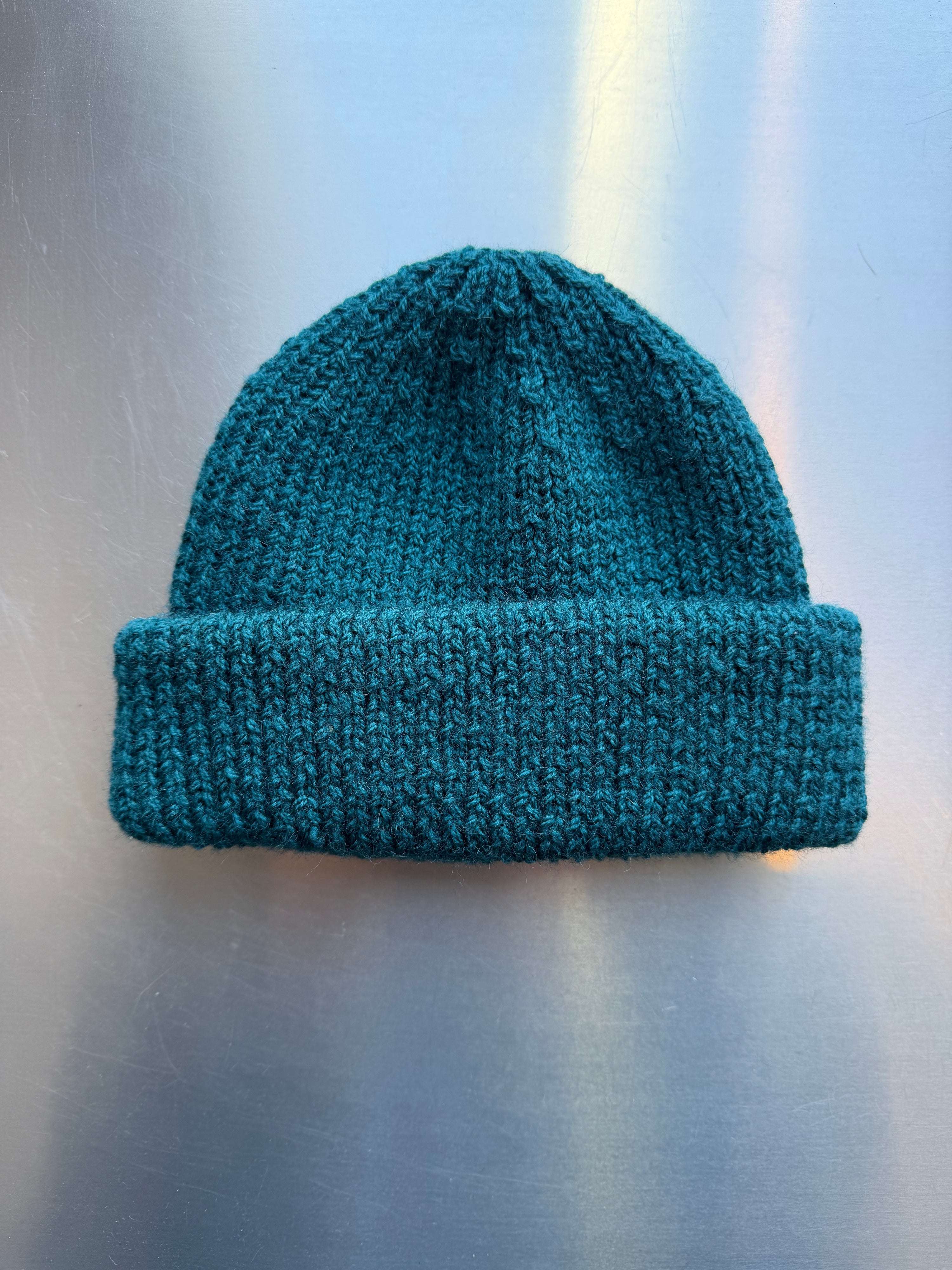 A teal coloured Cat Street hand-knit fishermans beanie with folded cuff, displayed on a brushed metal surface