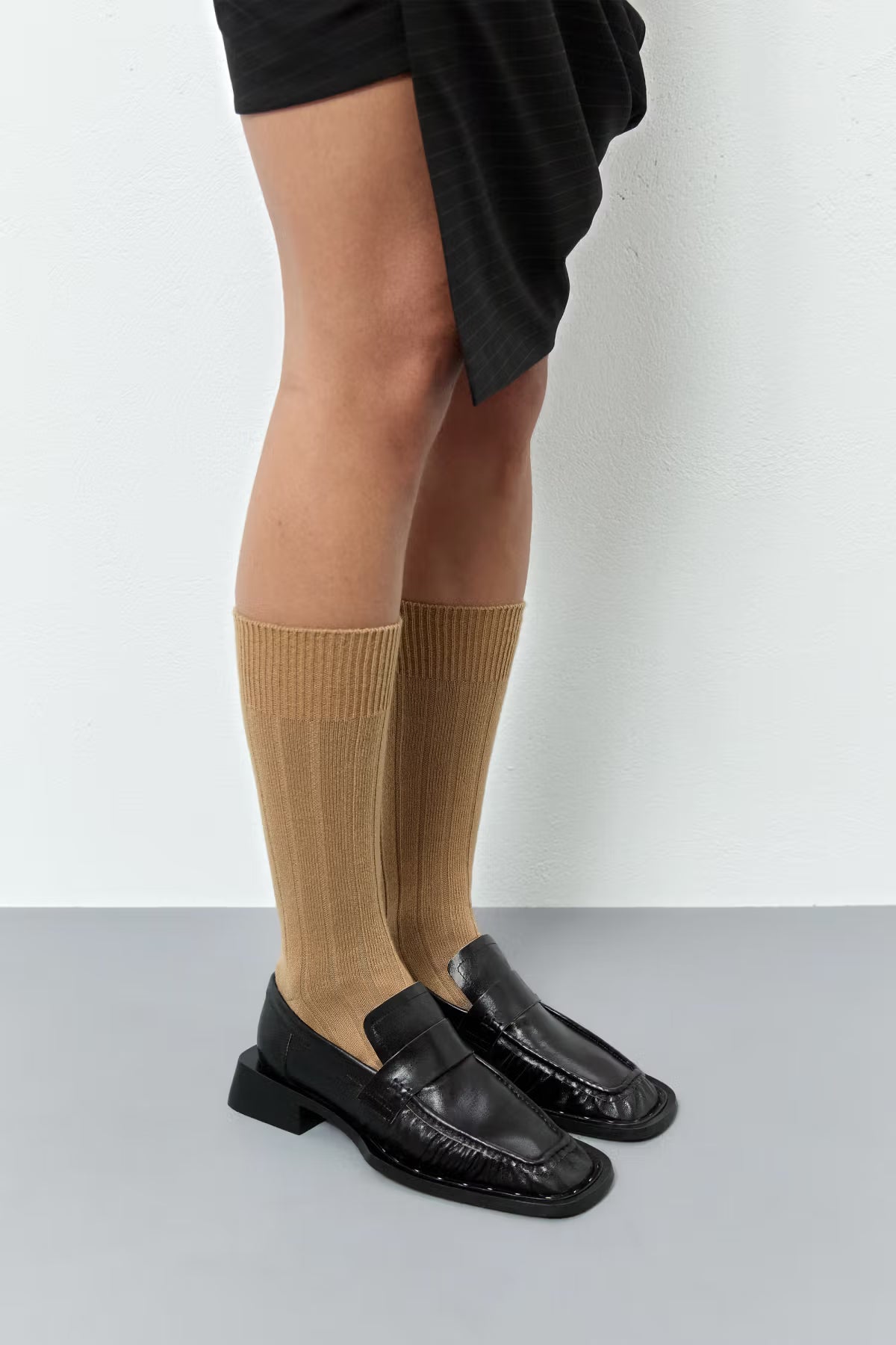Person wearing brown baige socks pulled up and black Miista Airi loafers on a light grey background