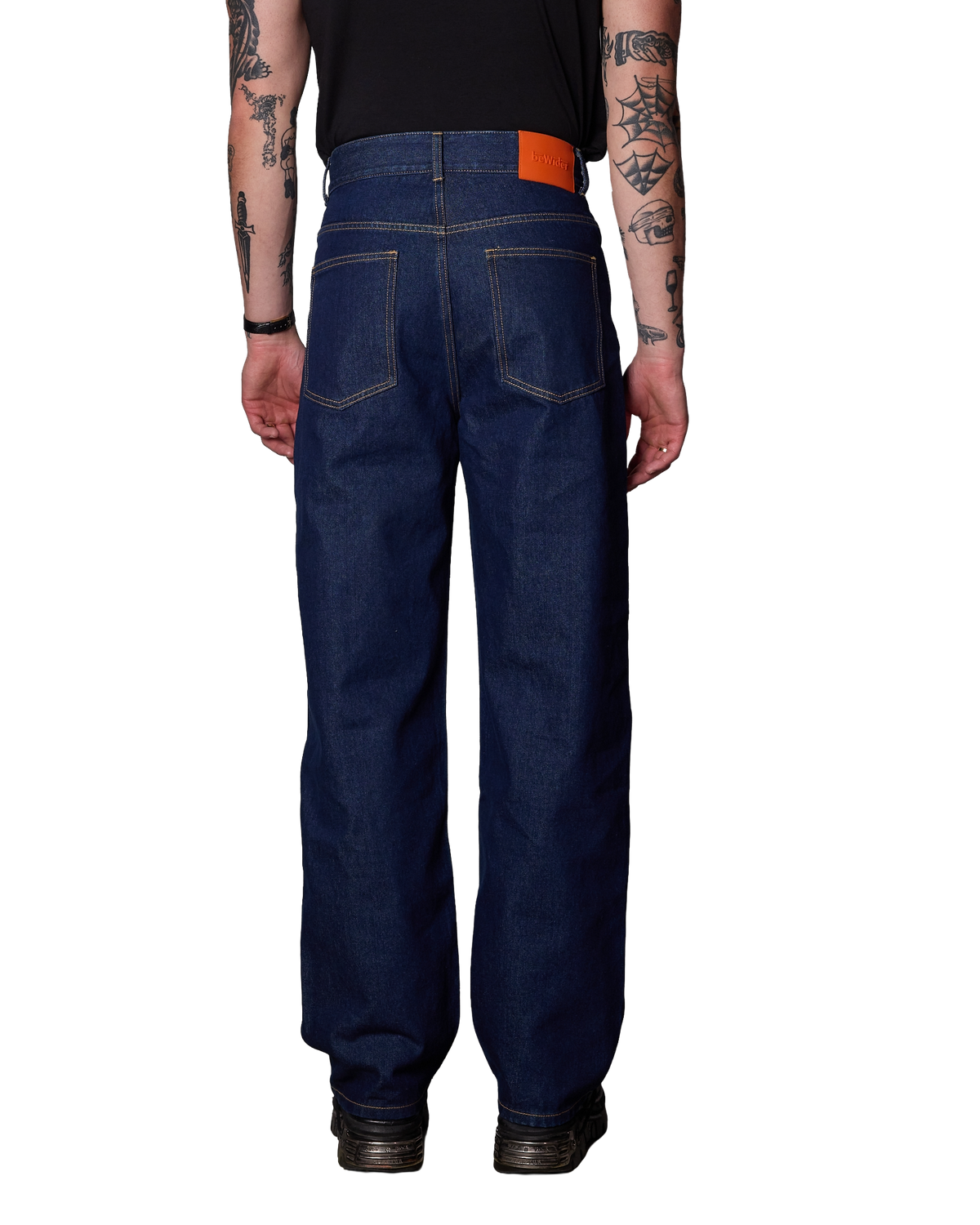 Rear view of a person wearing  bewider super loose dark blue denim jeans indigo with a bright orange brand patch, a black shirt, and black shoes against a plain white background.