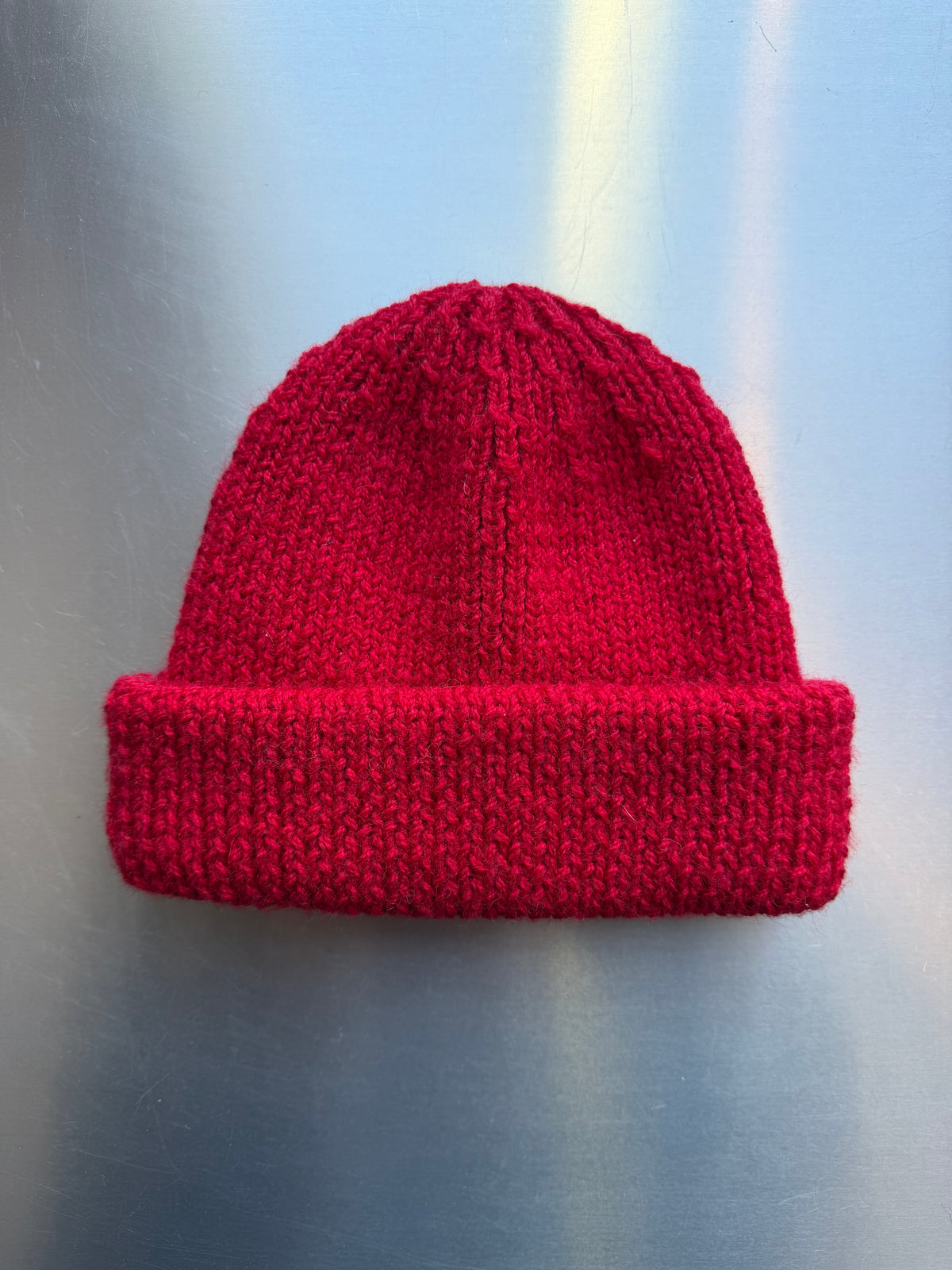 A Cat Street hand knitted fishermans beanie in raspberry red with folder cuff, displayed against a brushed metallic grey background.