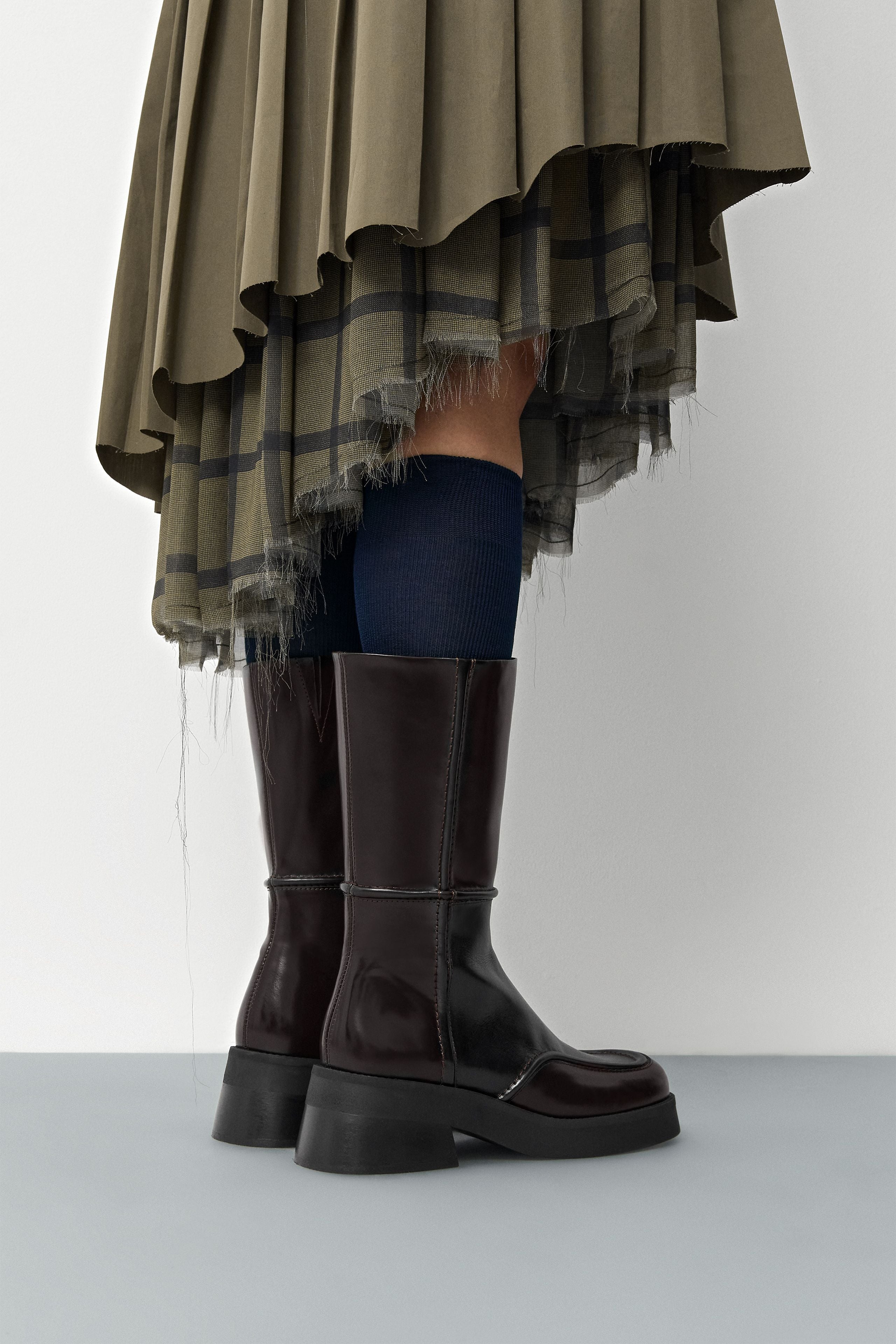 Person wearing a green plaid skirt and dark brown Miista Inne boots on a light gray background.